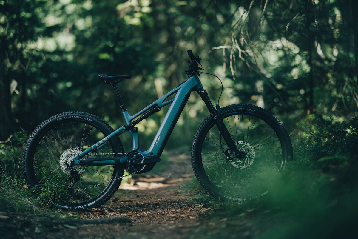 Ein blaues Cube Stereo Hybrid E-Mountainbike steht auf einem Waldtrail, umgeben von grünem Laub und weichem Licht.
