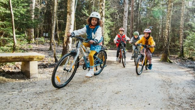 Kinder in bunten Jacken fahren auf Kinderfahrrädern durch einen Wald 